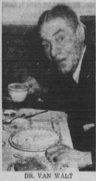 An older man with a slight smile is sitting at a dining table, holding a cup in one hand and gesturing with the other, surrounded by plates of food.